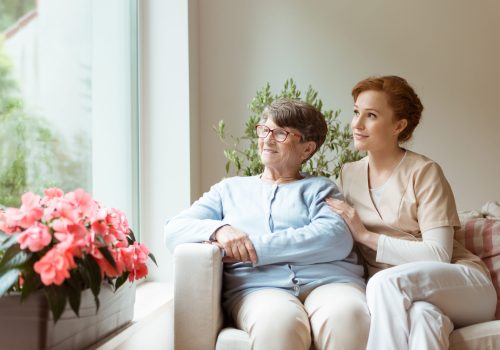 Geriatric woman and her professional caretaker sitting on a couch and looking through a window in a living room. Blooming flowers on a windowsill.